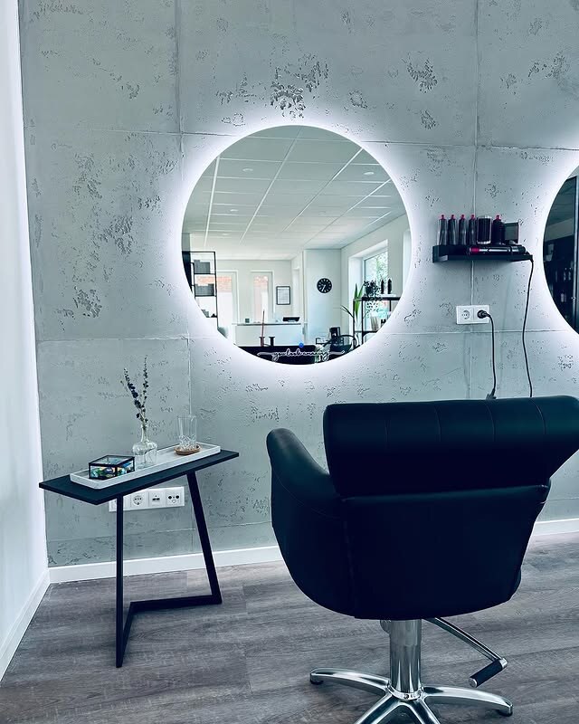 Arbeitsplatz mit rundem Leuchtspiegel und schwarzem Friseurstuhl vor einer Betonwand